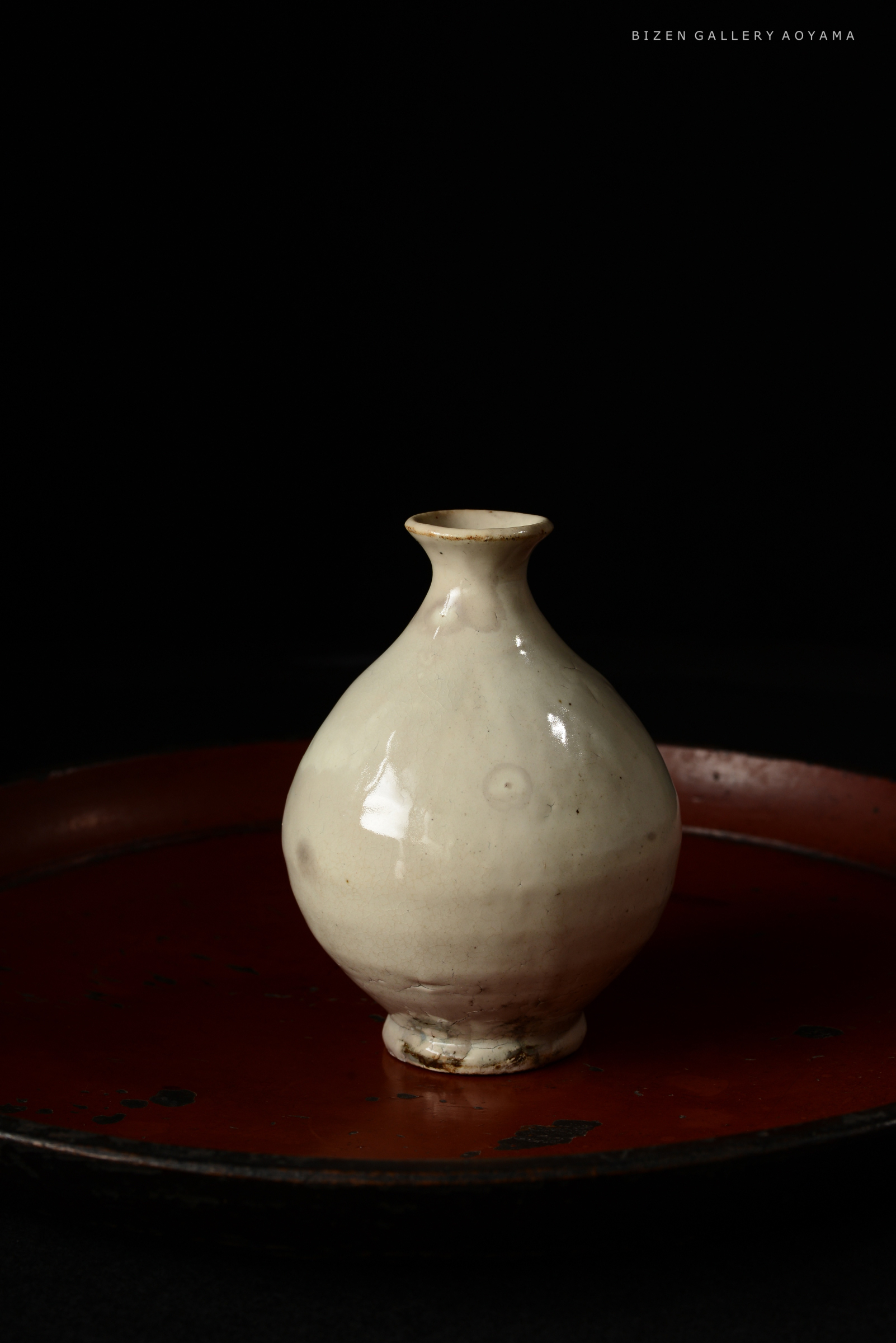 A small, white ceramic vase with a rounded body and narrow neck, displayed on a circular red tray against a black background.