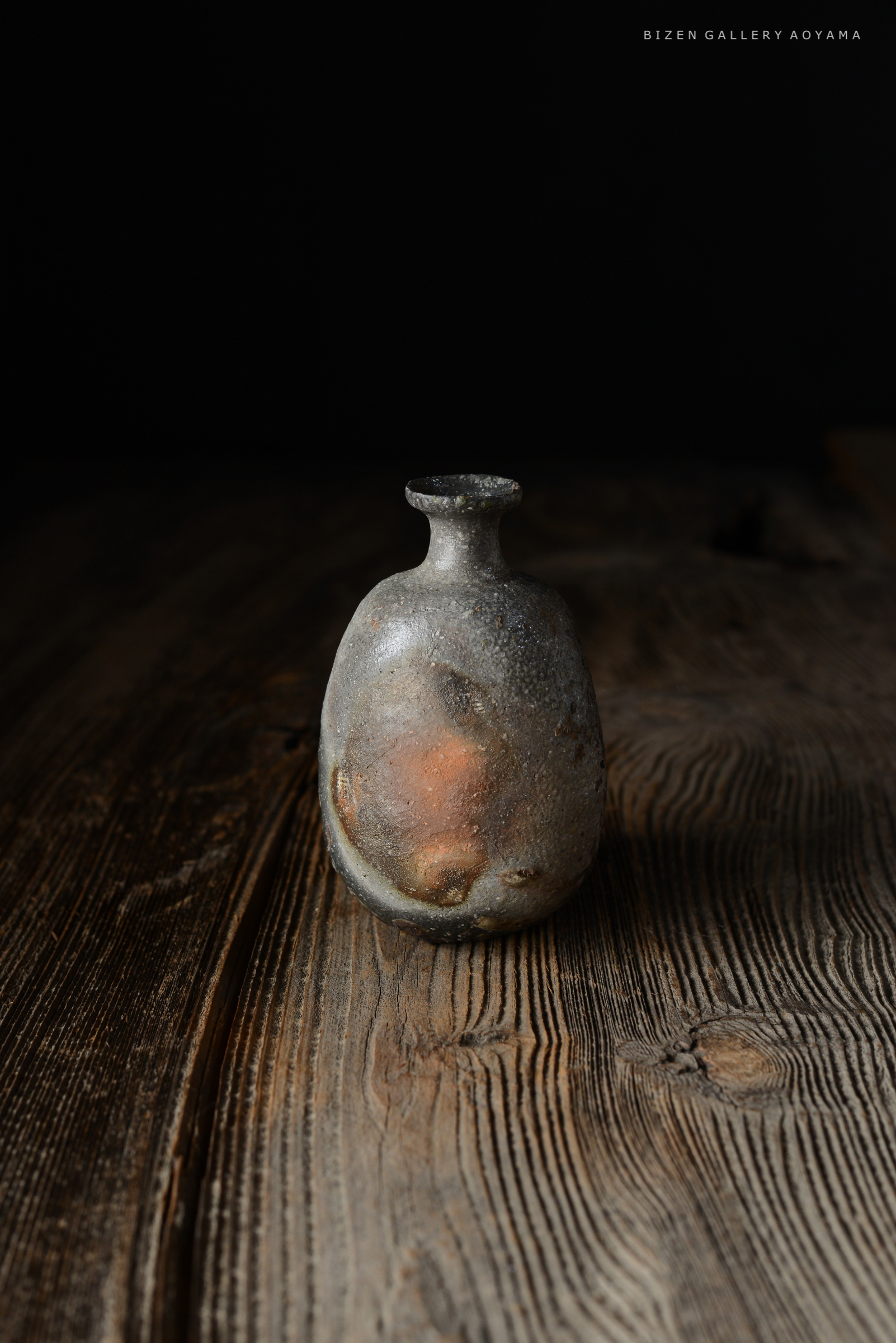 A small, textured ceramic vase with an elongated neck, featuring a mix of gray and earthy tones, placed on a rustic wooden surface.
