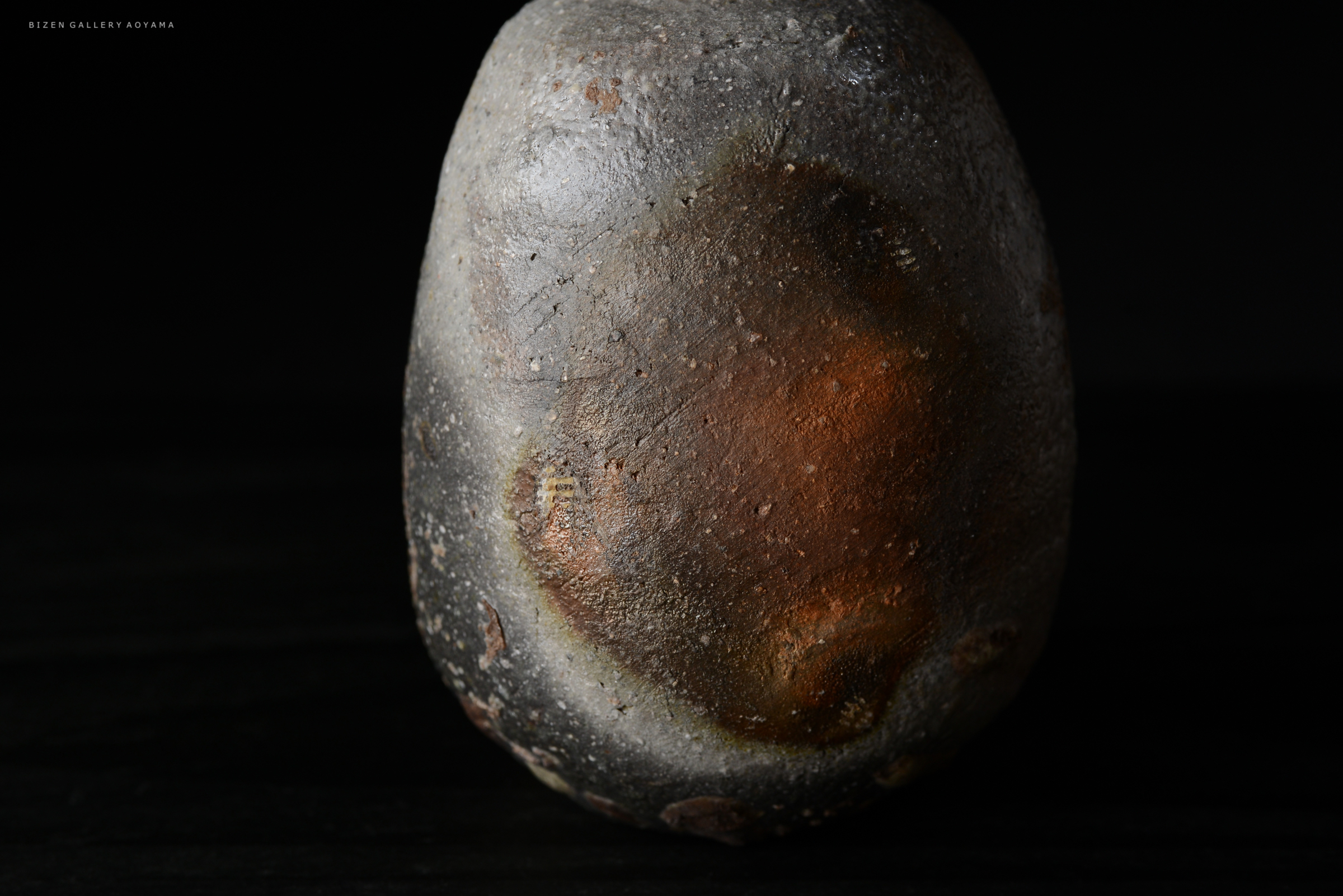 A close-up view of a textured, rounded ceramic piece with earthy tones, featuring a smooth surface and subtle color variations against a dark background.