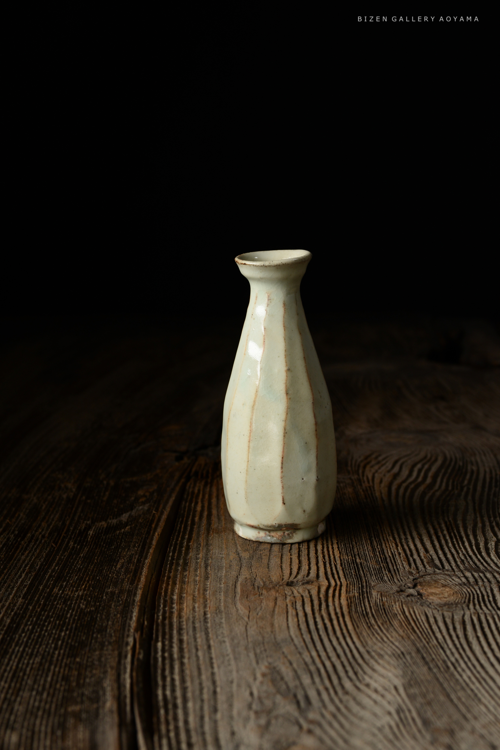A small, pale ceramic vase with vertical stripes, placed on a textured wooden surface against a dark background.