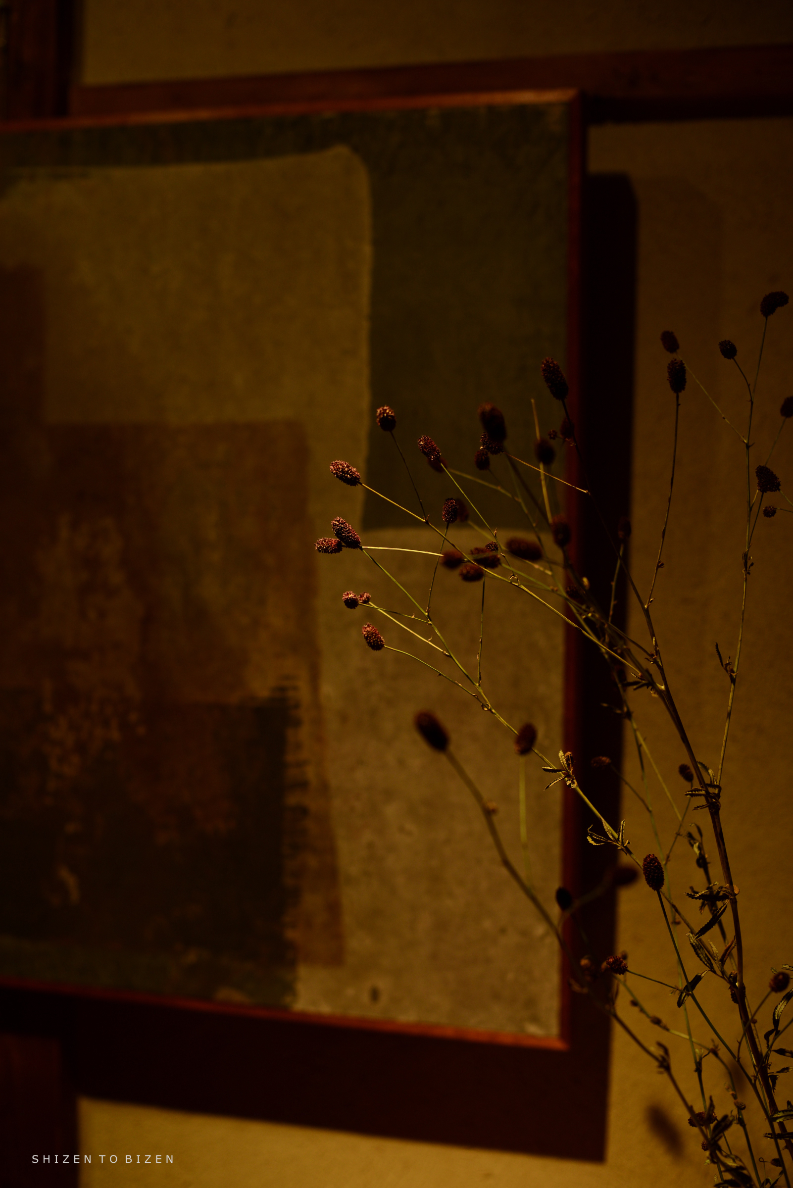 A minimalist still life featuring a dried branch with small buds in the foreground, set against a textured abstract background with muted colors.