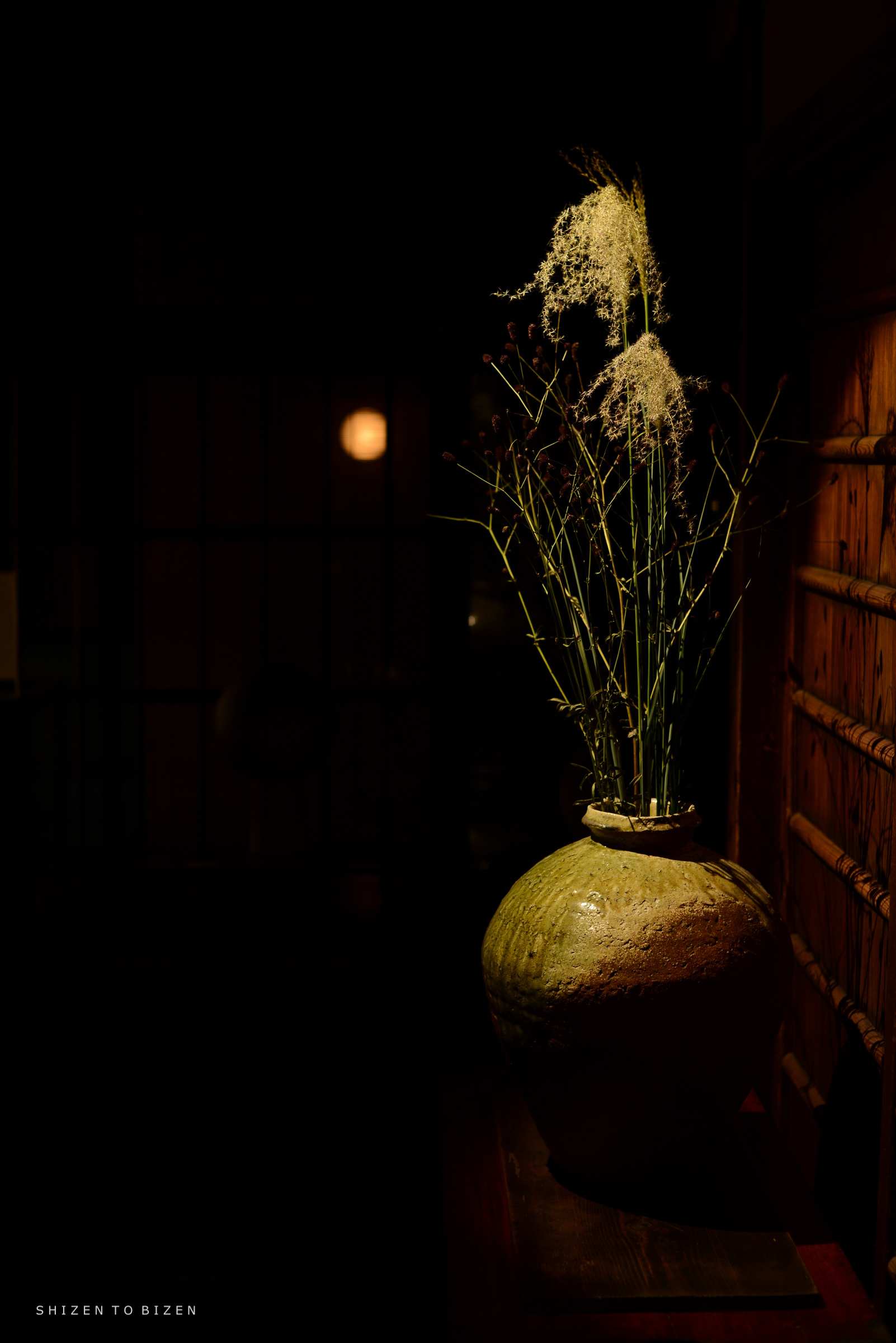 A ceramic vase with dried plants and flowers against a dark background, softly illuminated by ambient light.