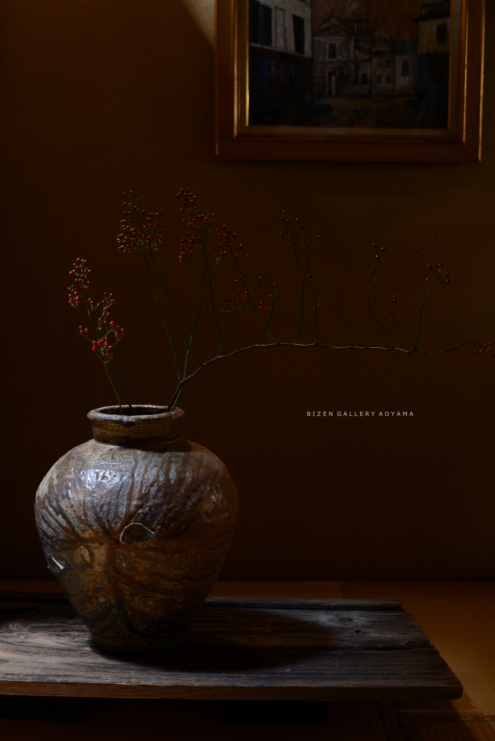 A decorative Bizen pottery vase displaying slender branches with small red berries, set on a rustic wooden surface with warm ambient lighting.