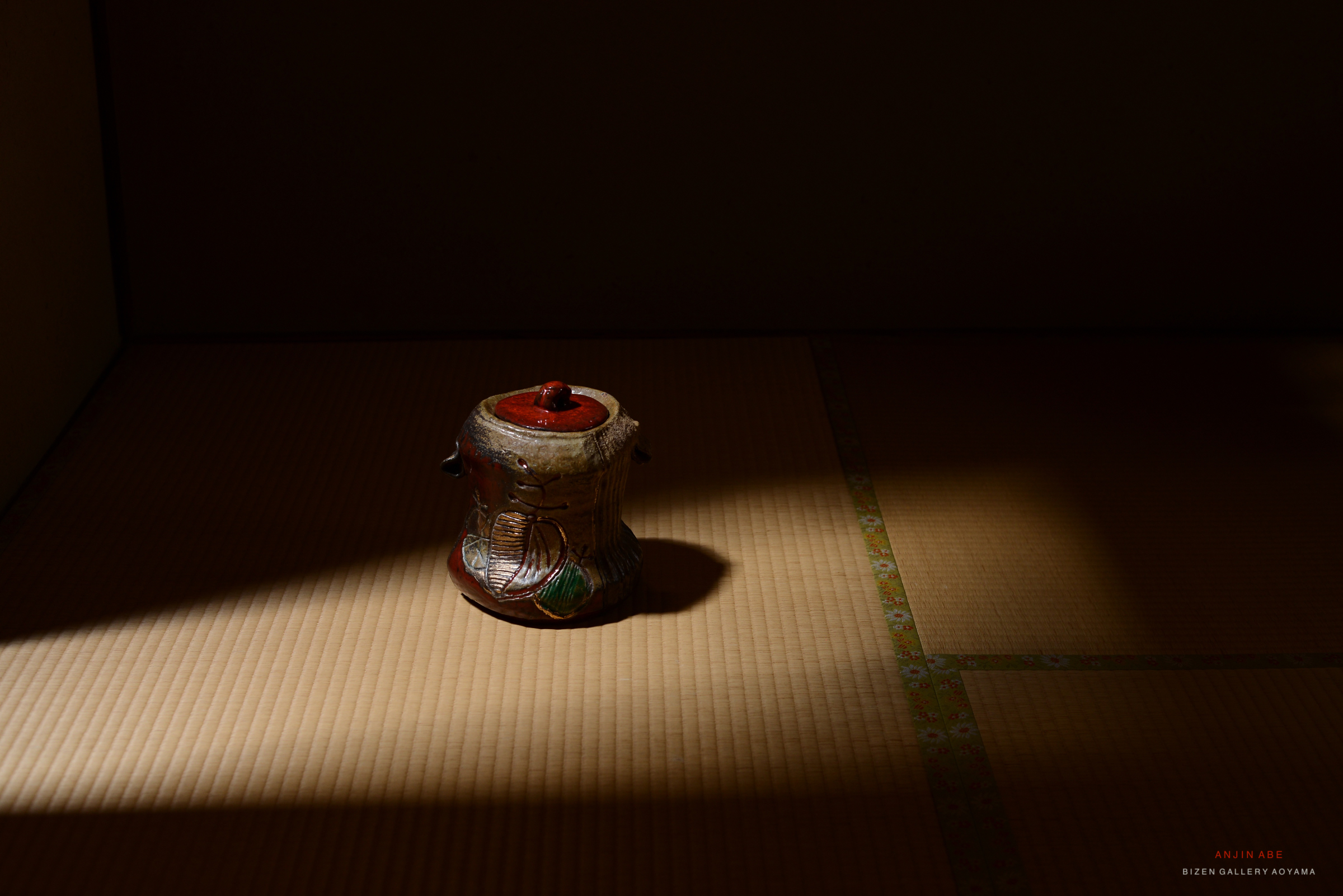 A decorative ceramic jar with a red lid, placed on a tatami mat, illuminated by soft natural light.