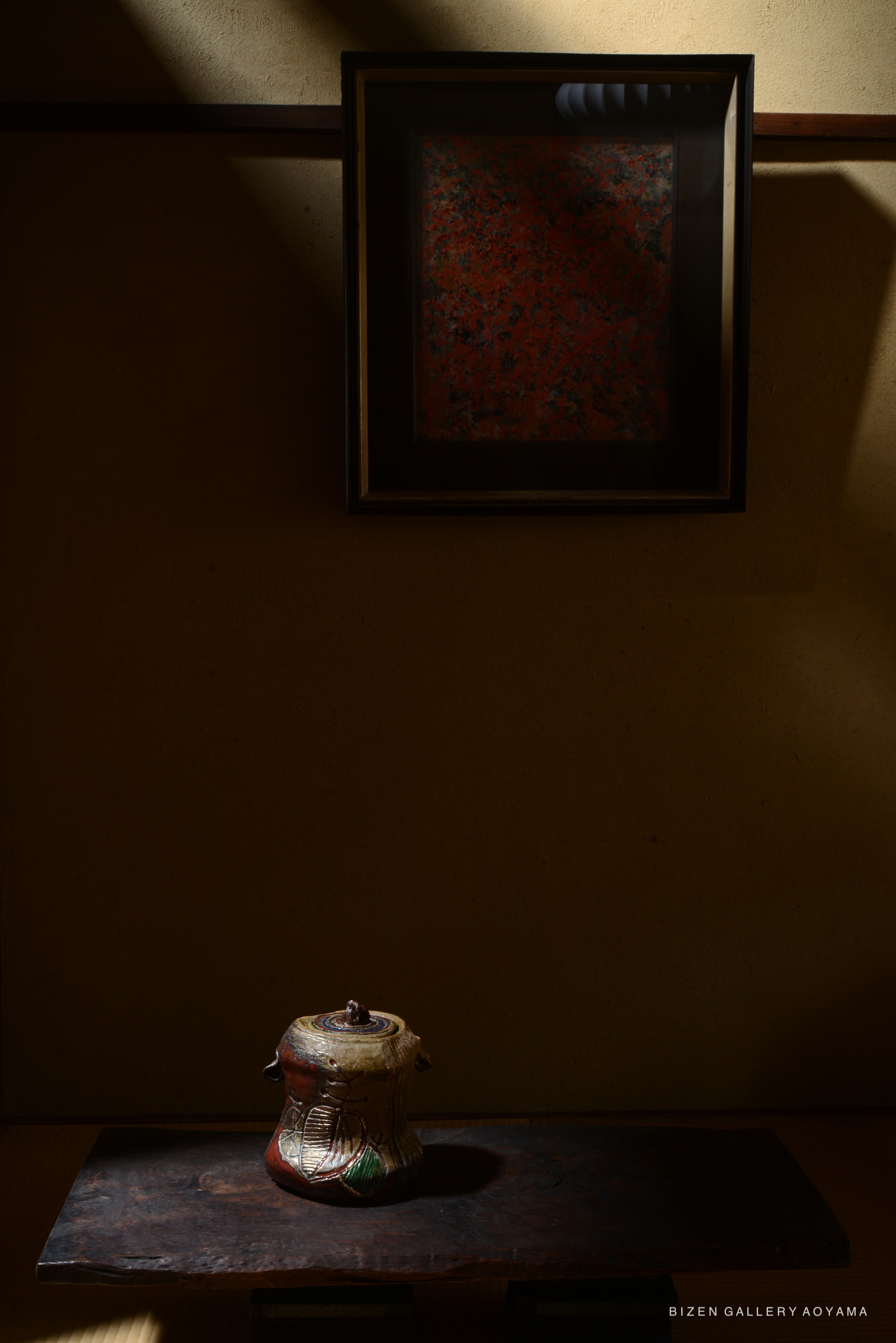 A rustic ceramic teapot sits on a wooden surface in a dimly lit room, with a framed artwork partially illuminated on the wall above it.