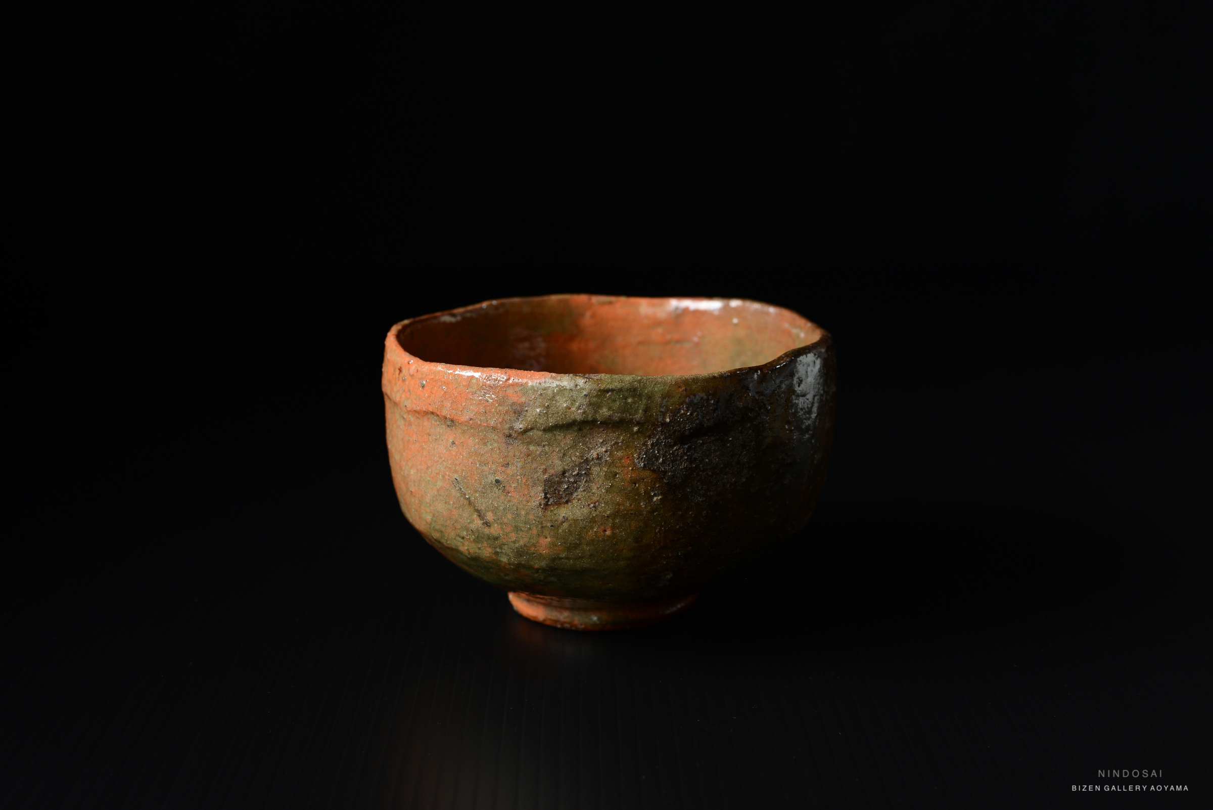 A handcrafted clay bowl with a rough texture and an irregular shape, featuring earthy orange and green tones, set against a dark background.