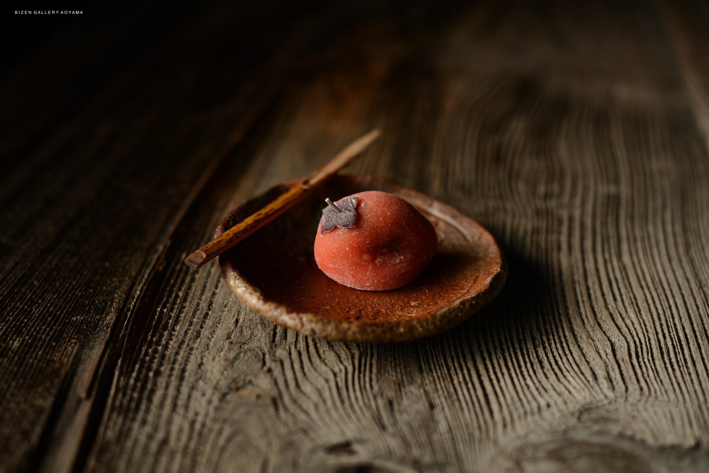 A small round dessert resembling a persimmon, placed on a rustic brown plate, accompanied by a piece of cinnamon stick, set against a wooden surface.